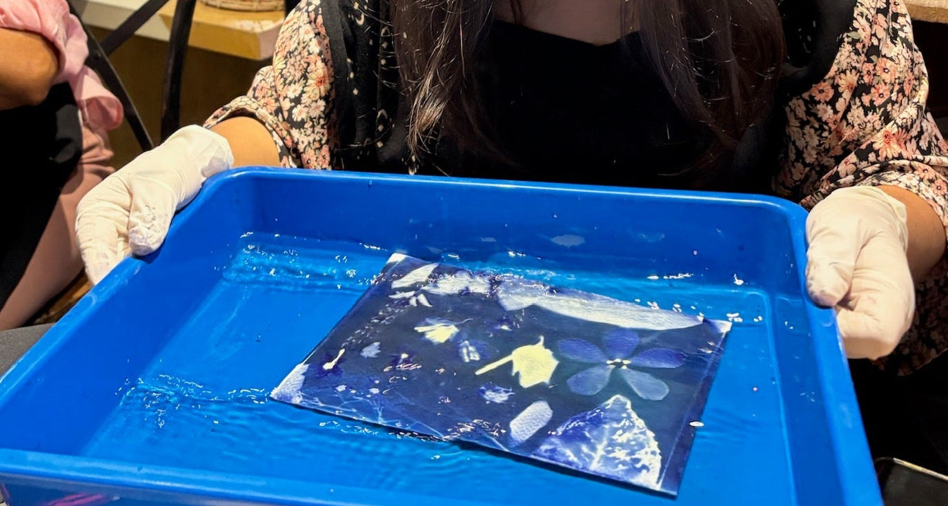 Person wearing gloves and holding a blue tray with a small painting inside, in an indoor setting.
