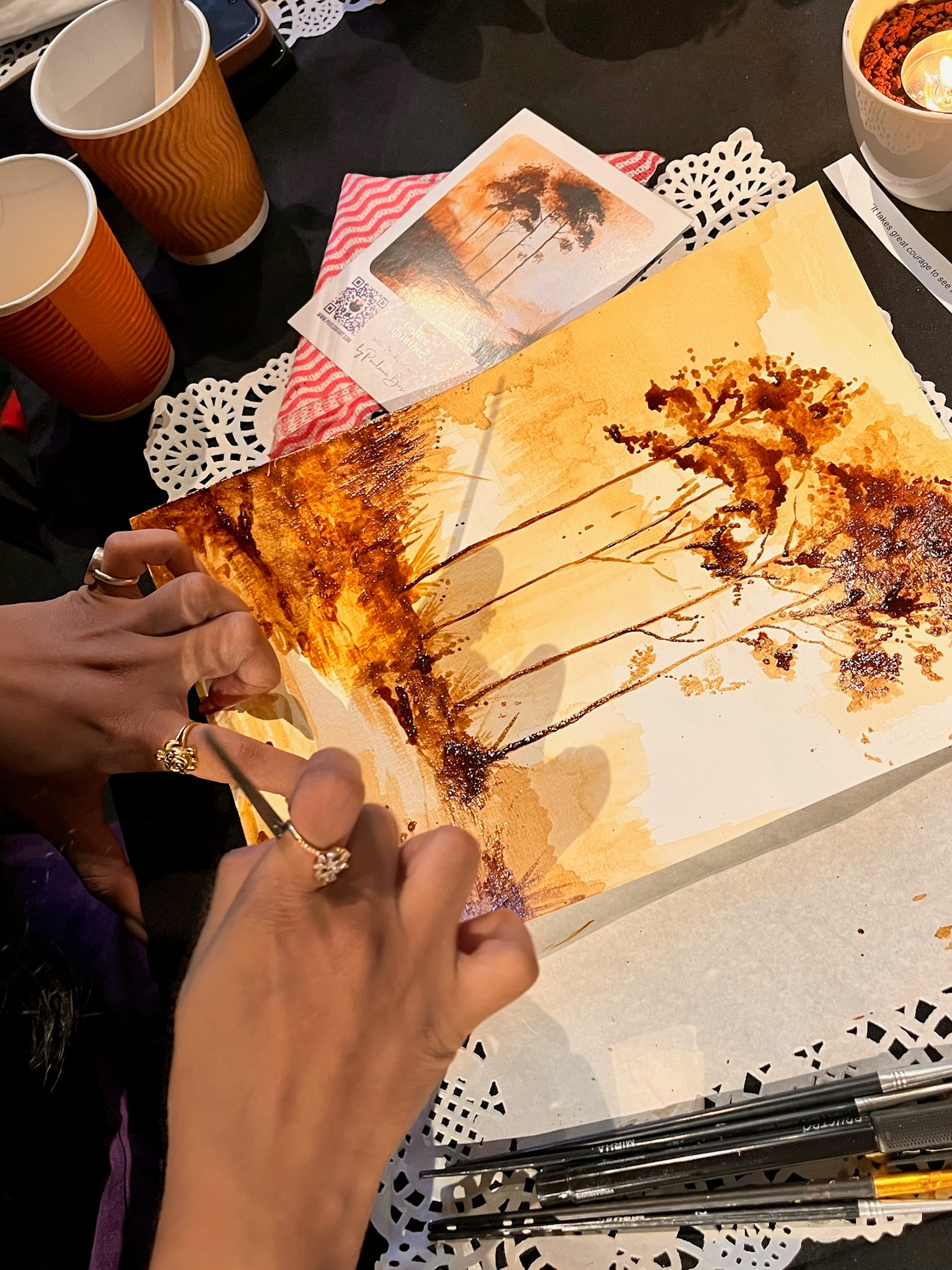Artistic coffee painting on a textured surface with hands holding it, surrounded by cups and stationery.